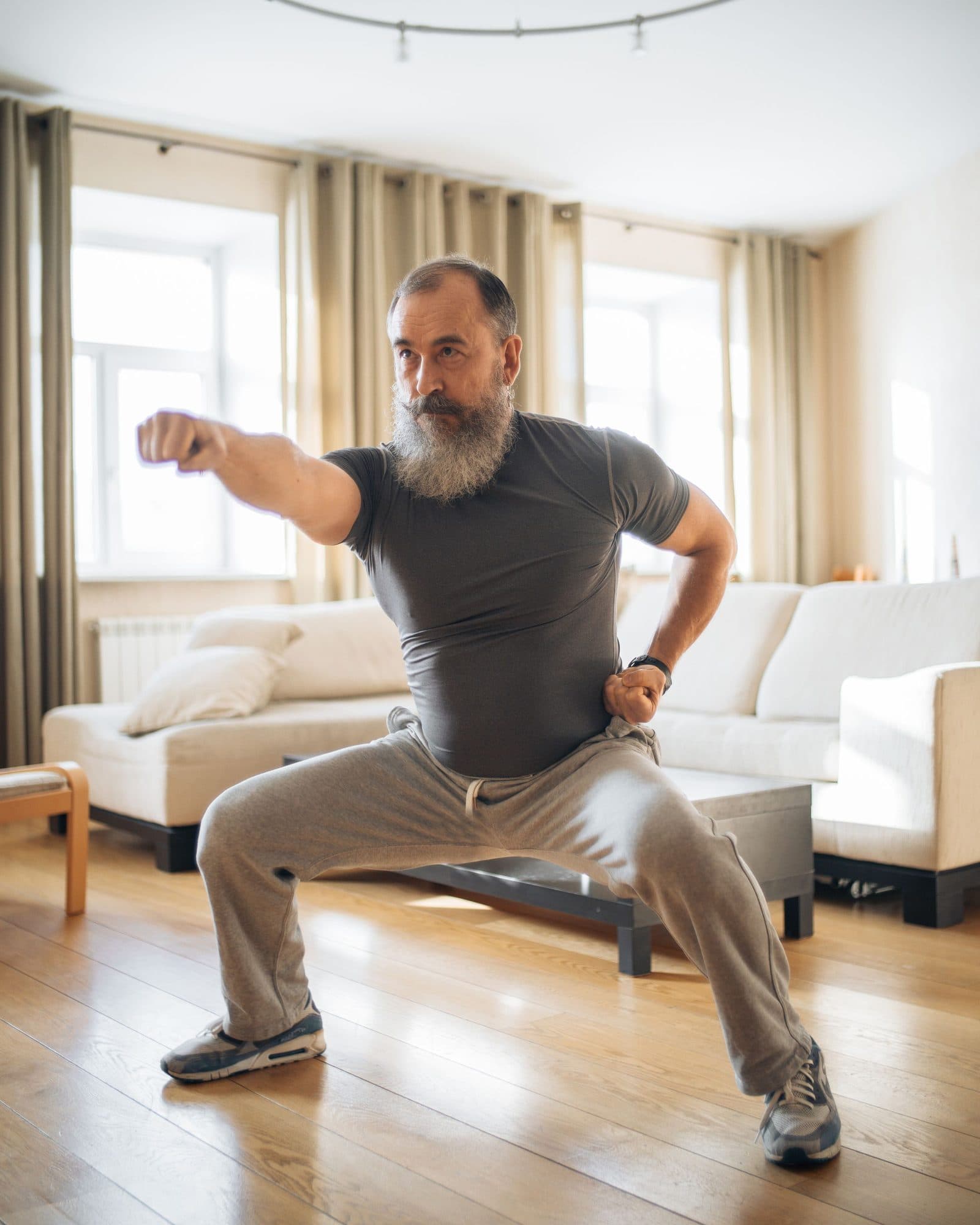 An older man holding a deep squat in his sunlit living room, one arm extended.